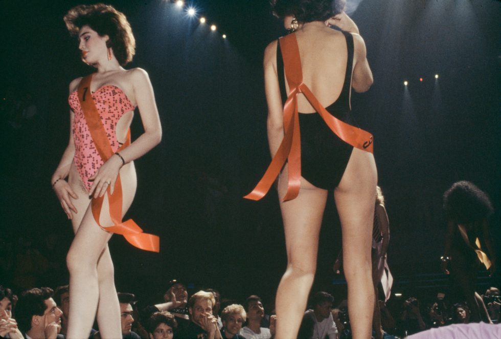 Two light-skinned individuals in swimsuits and orange sashes stand on a stage facing opposite directions before an audience under spotlights.