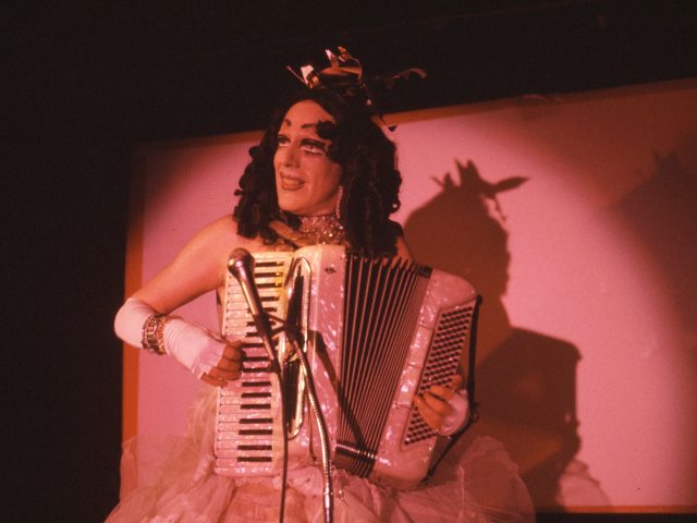 A color photograph of a a performer on stage in pink lighting.