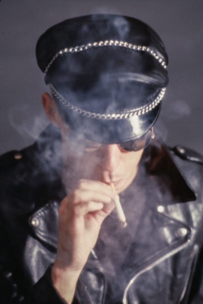 A man in a black leather cap and jacket, wearing sunglasses, is smoking a cigarette, with smoke swirling around him against a dark grey