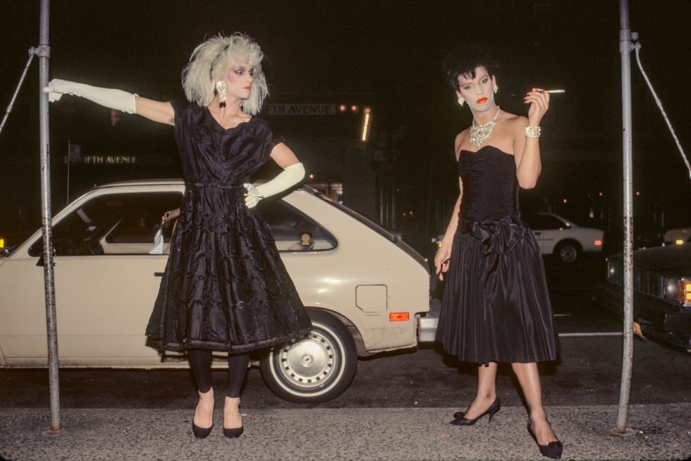 Two elegantly dressed individuals in black gowns pose dramatically on a city sidewalk at night, one holding a cigarette and the other extending a gloved arm.