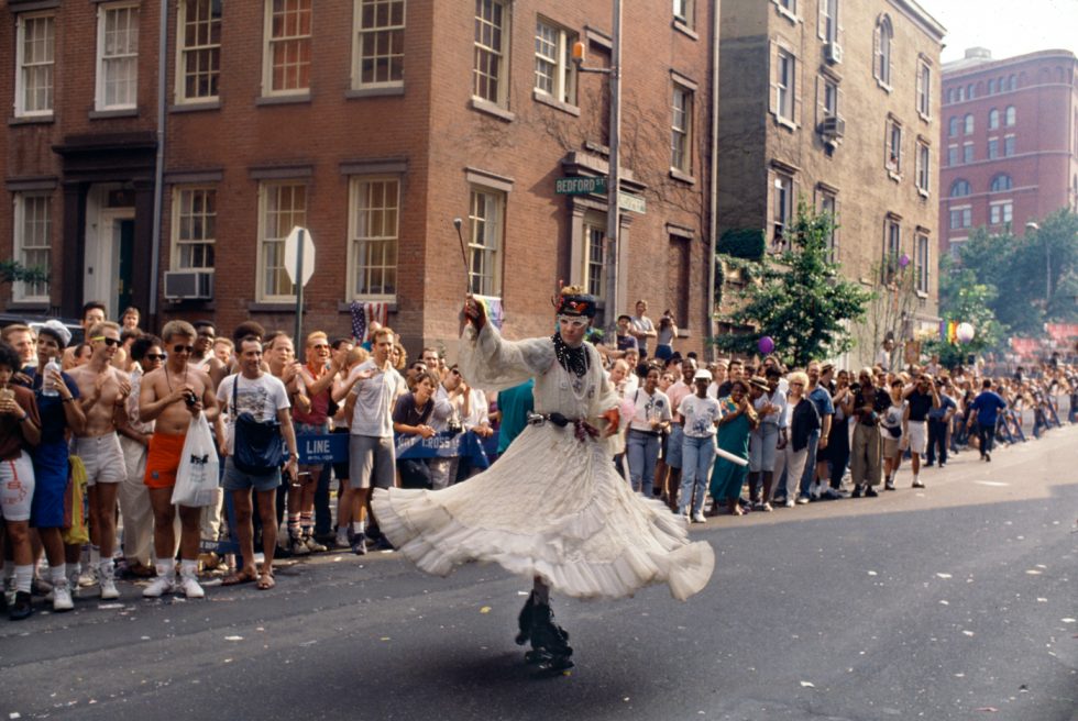 A color photograph of a pride parade with a person in roller skates and a white dress.