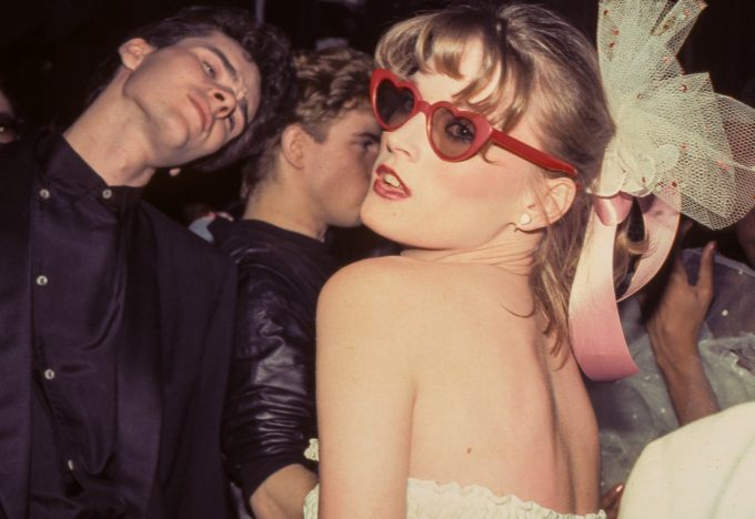 A woman in heart-shaped sunglasses and a large bow looks over her shoulder at a crowded nightclub, surrounded by stylishly dressed partygoers.