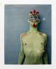 Person with green-painted body and flower-like face paint stands against a blue background with a neutral expression.