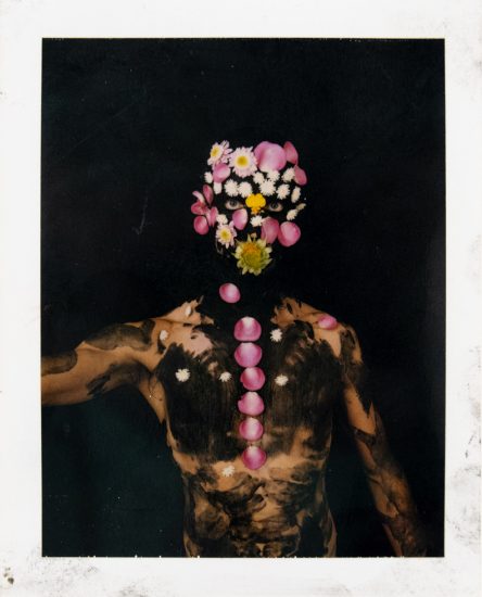 A shirtless person with dark body paint stands against a black background, their face and spine adorned with pink petals and small white flowers.