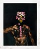 A shirtless person with dark body paint stands against a black background, their face and spine adorned with pink petals and small white flowers.