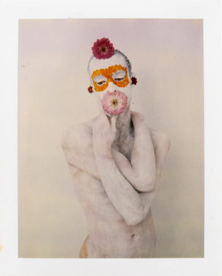 Figure covered in white with a vibrant floral mask (orange eyes, pink mouth, red accents) against a plain backdrop.