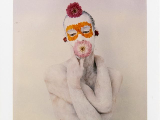 Figure covered in white with a vibrant floral mask (orange eyes, pink mouth, red accents) against a plain backdrop.