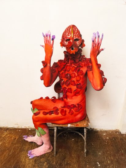 Red-painted figure with petal mask and adornments sits on a chair against a white wall, raising purple-tipped hands and feet.