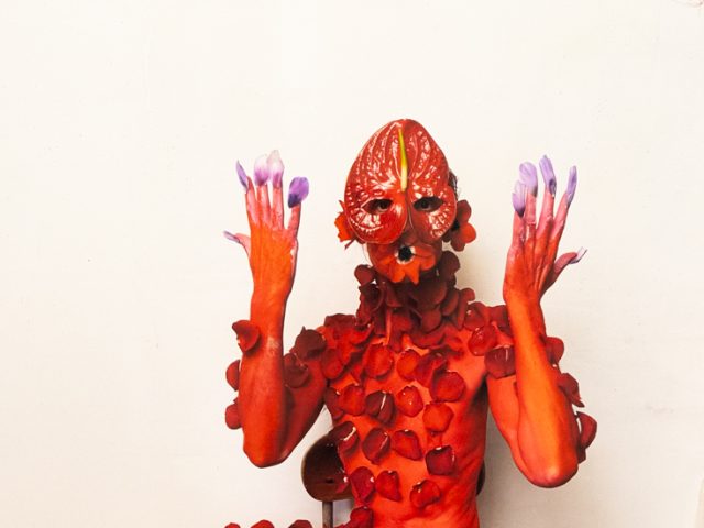 Red-painted figure with petal mask and adornments sits on a chair against a white wall, raising purple-tipped hands and feet.