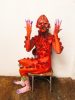 Red-painted figure with petal mask and adornments sits on a chair against a white wall, raising purple-tipped hands and feet.