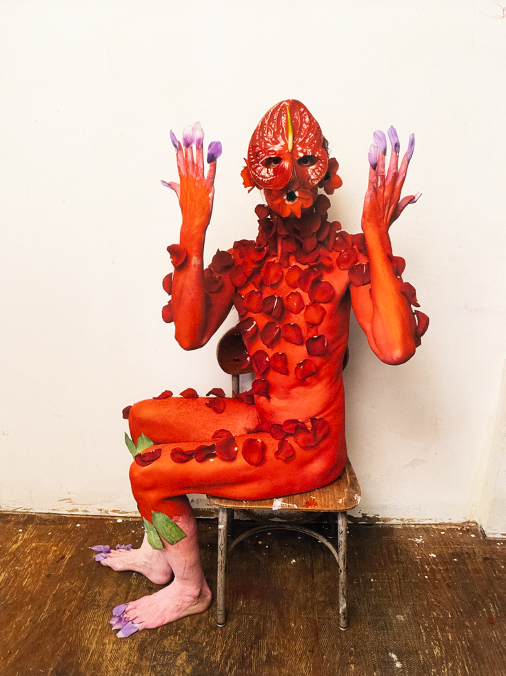 Red-painted figure with petal mask and adornments sits on a chair against a white wall, raising purple-tipped hands and feet.