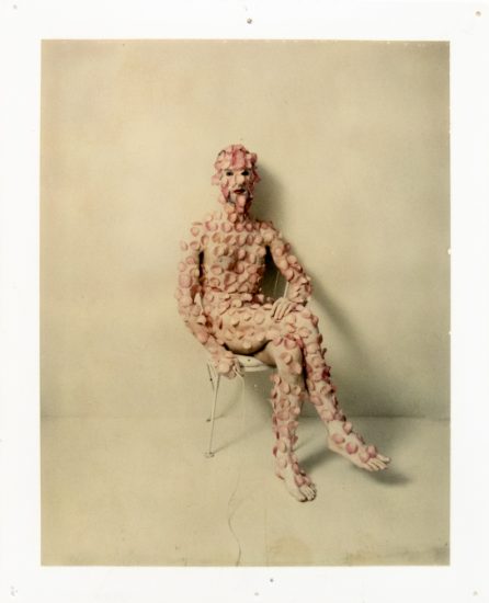 A figure densely covered in pale pink discs sits on a white chair against a pale wall, obscuring their features.