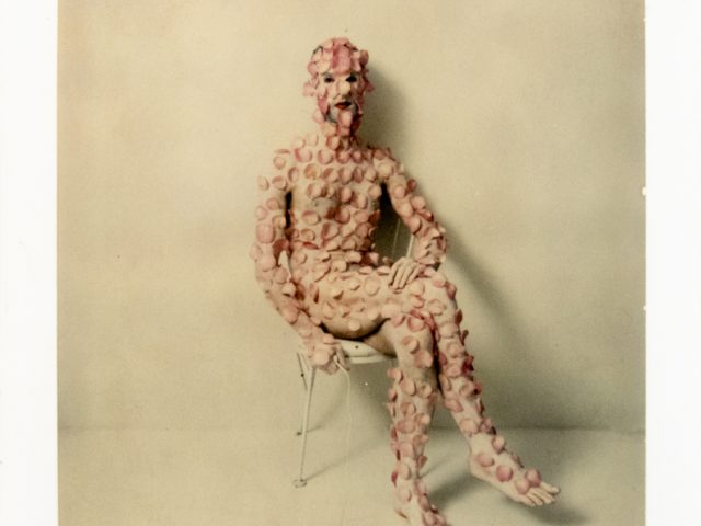 A figure densely covered in pale pink discs sits on a white chair against a pale wall, obscuring their features.