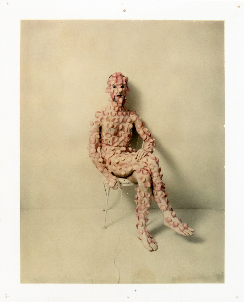 A figure densely covered in pale pink discs sits on a white chair against a pale wall, obscuring their features.