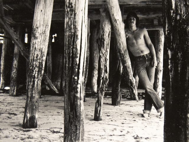 A black and white photograph of a shirtless man standing under a dock.