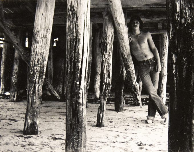 A black and white photograph of a shirtless man standing under a dock.
