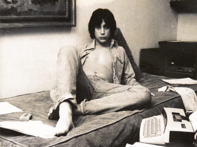 A black and white photograph of a young man sitting on a bed.
