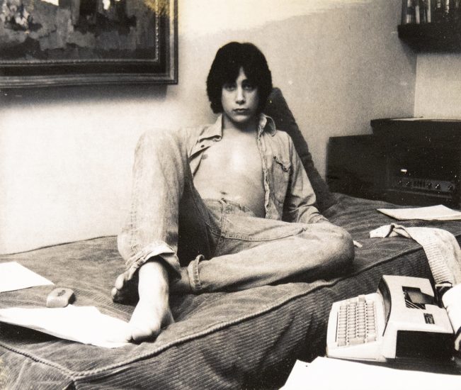 A black and white photograph of a young man sitting on a bed.