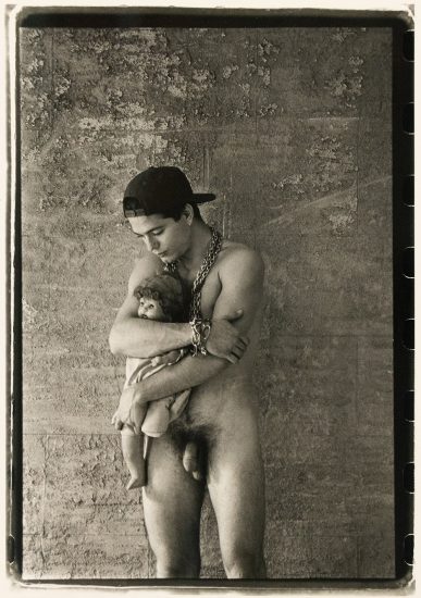 Nude man in a backward cap holds a doll tenderly against a textured wall, wearing heavy chain jewelry.