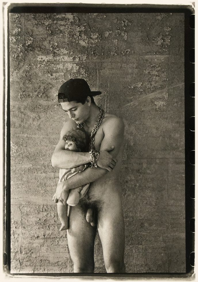 Nude man in a backward cap holds a doll tenderly against a textured wall, wearing heavy chain jewelry.