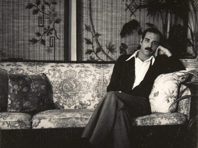 A man with a mustache lounges on an ornate floral-patterned sofa in a vintage room with leafy wallpaper and potted plants.