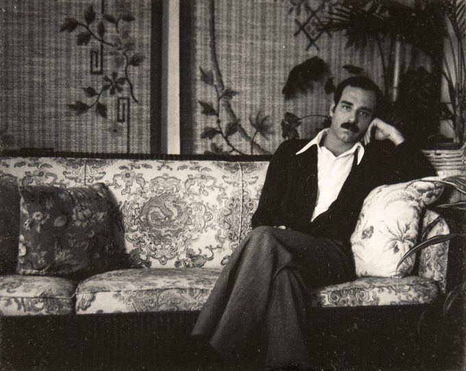 A man with a mustache lounges on an ornate floral-patterned sofa in a vintage room with leafy wallpaper and potted plants.