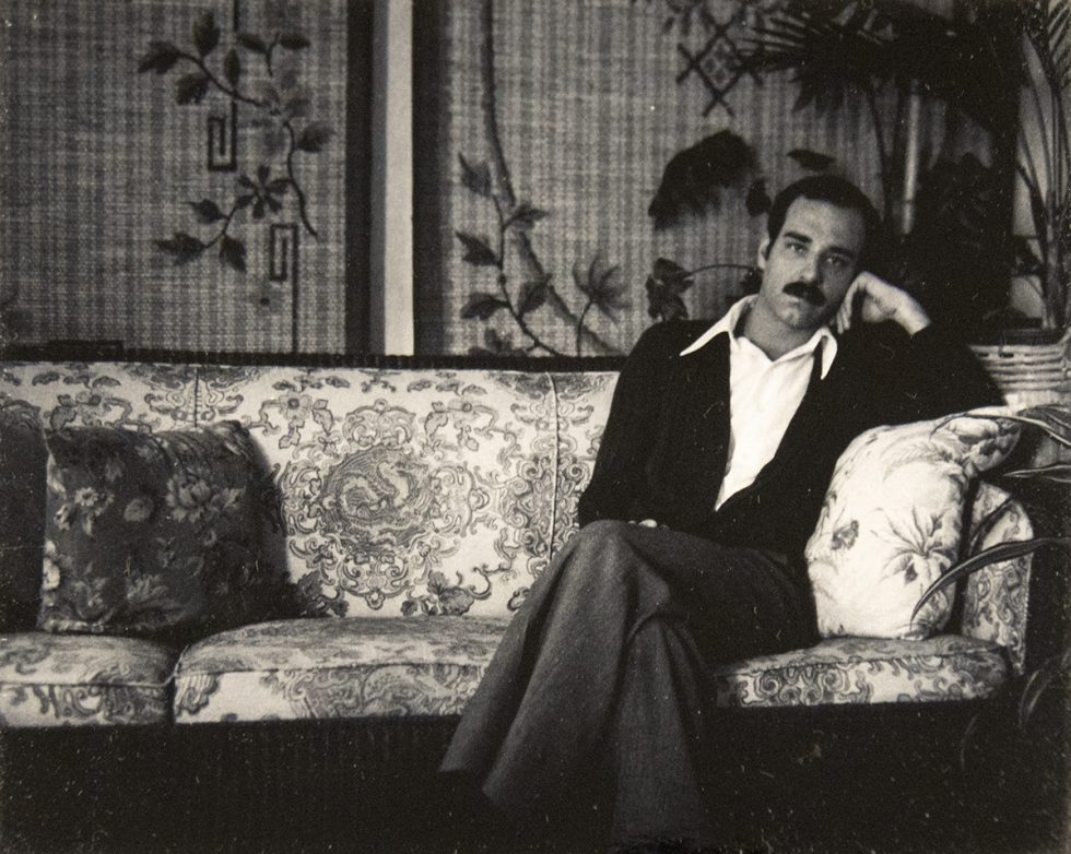 A man with a mustache lounges on an ornate floral-patterned sofa in a vintage room with leafy wallpaper and potted plants.