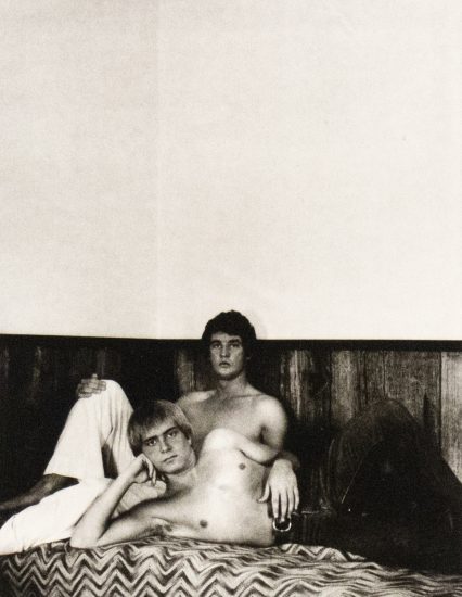 A black and white photograph of two men shirtless in bed.