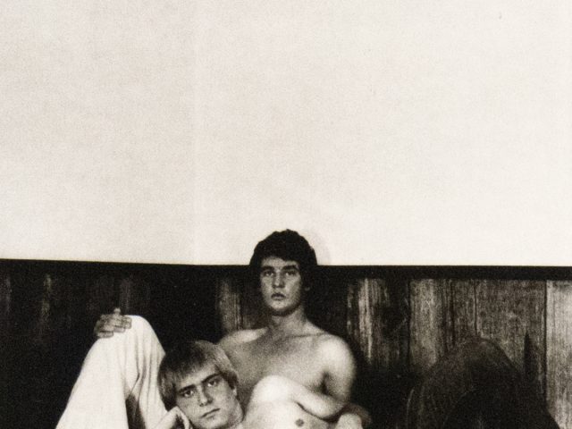 A black and white photograph of two men shirtless in bed.