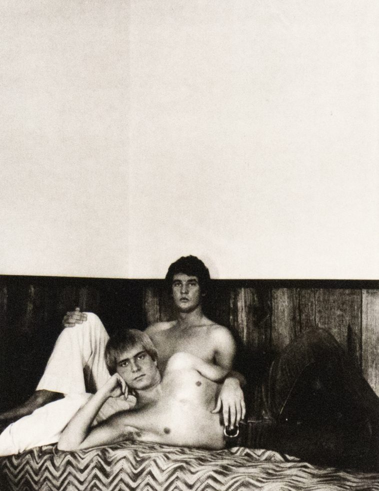 A black and white photograph of two men shirtless in bed.