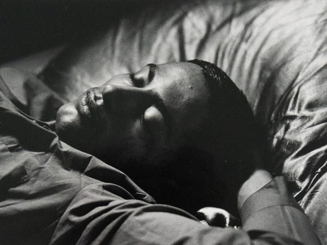 A black and white close-up photograph of a man sleeping in bed. His eye are closed and his expression is soft.