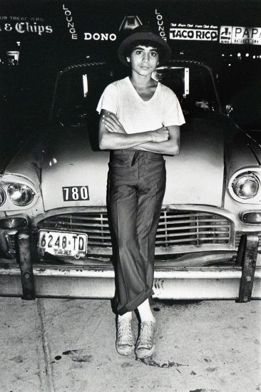 A boy in a hat, t-shirt, and trousers stands with crossed arms in front of a parked taxi with the license plate "6248-TD". Neon signs for "Lounge" and "Taco Rico" are visible in the background.