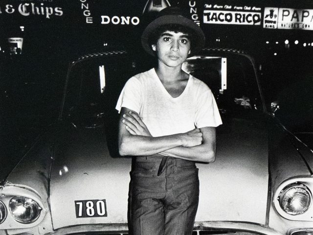 A boy in a hat, t-shirt, and trousers stands with crossed arms in front of a parked taxi with the license plate "6248-TD". Neon signs for "Lounge" and "Taco Rico" are visible in the background.