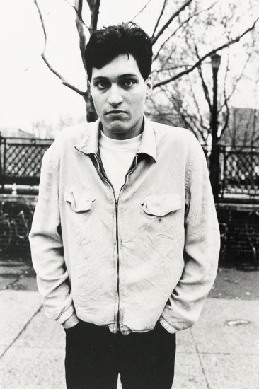 A young man in a light zip-up jacket stands against bare winter trees.