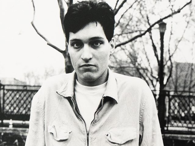 A young man in a light zip-up jacket stands against bare winter trees.