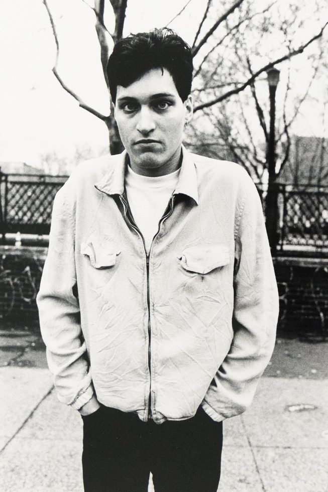 A young man in a light zip-up jacket stands against bare winter trees.