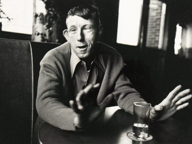 A black and white photograph captures a man seated at a table, gesturing with both hands while looking directly at the viewer. A glass with liquid rests on a coaster in the foreground.