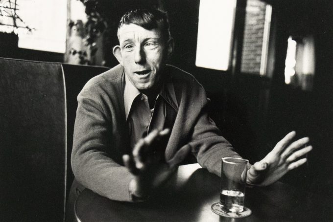 A black and white photograph captures a man seated at a table, gesturing with both hands while looking directly at the viewer. A glass with liquid rests on a coaster in the foreground.