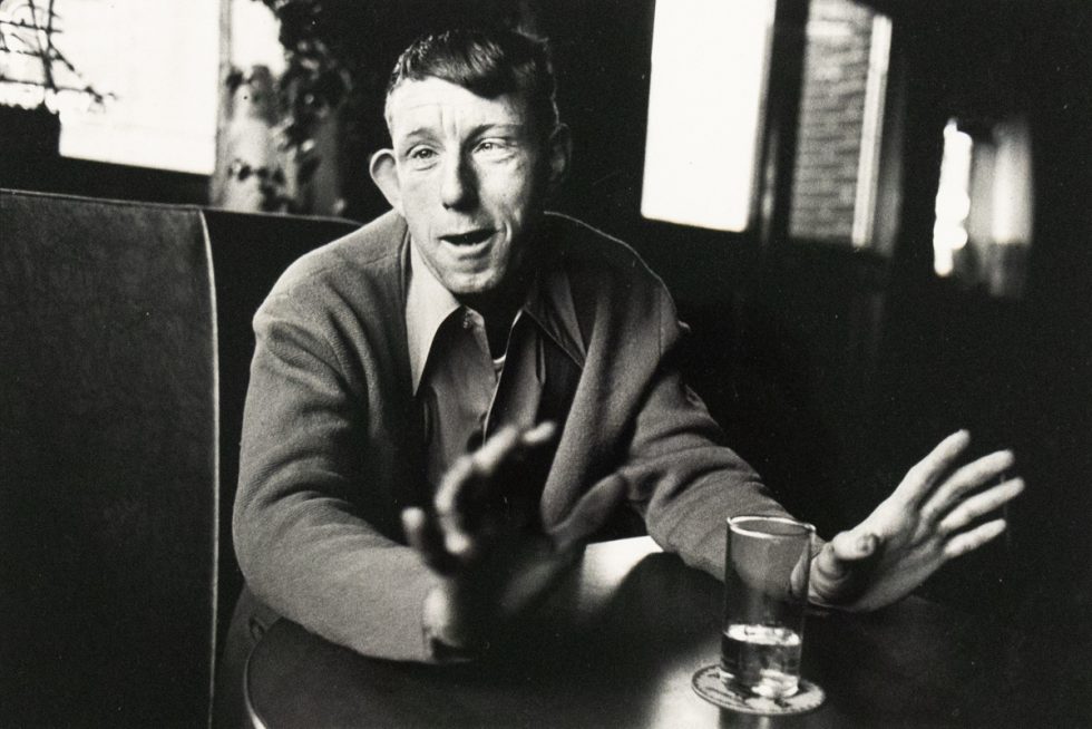 A black and white photograph captures a man seated at a table, gesturing with both hands while looking directly at the viewer. A glass with liquid rests on a coaster in the foreground.