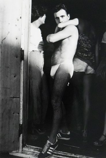 A shirtless man in a speedo stands in a doorway, looking over his shoulder at the viewer. Another person in striped pants stands next to him, facing away.