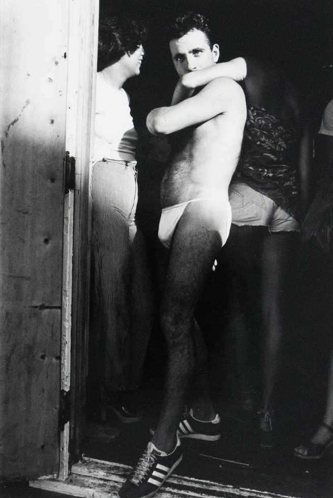 A shirtless man in a speedo stands in a doorway, looking over his shoulder at the viewer. Another person in striped pants stands next to him, facing away.