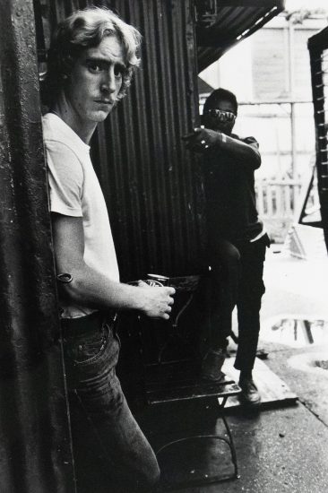 A blond man in a white t-shirt and jeans stands facing the viewer with an expression of mild surprise, holding a can. Behind him, another figure in sunglasses is partially visible pointing at the subject.