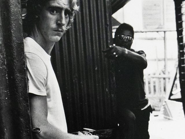 A blond man in a white t-shirt and jeans stands facing the viewer with an expression of mild surprise, holding a can. Behind him, another figure in sunglasses is partially visible pointing at the subject.