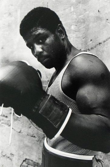A boxer wearing a mesh tank top and gloves poses with his fists raised, sweat visible on his face and arms, standing against a rough-textured wall.