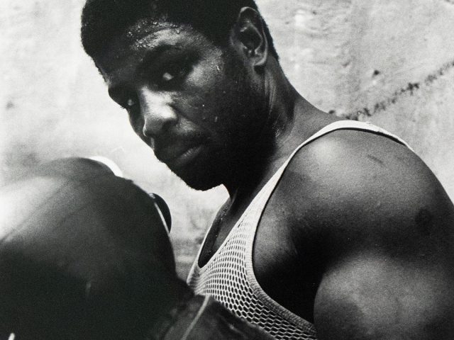 A boxer wearing a mesh tank top and gloves poses with his fists raised, sweat visible on his face and arms, standing against a rough-textured wall.