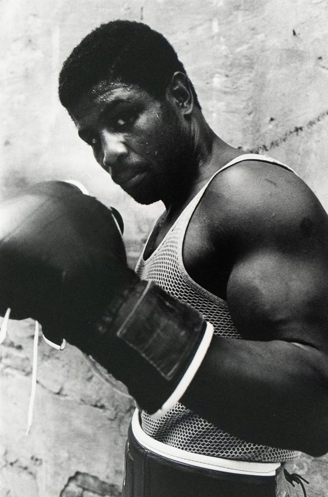 A boxer wearing a mesh tank top and gloves poses with his fists raised, sweat visible on his face and arms, standing against a rough-textured wall.