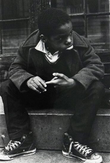 A black and white photograph captures a young boy sitting on a concrete stoop, looking towards the right side of the frame. He wears a dark coat over a collared shirt and v-neck sweater, with his hands clasped.