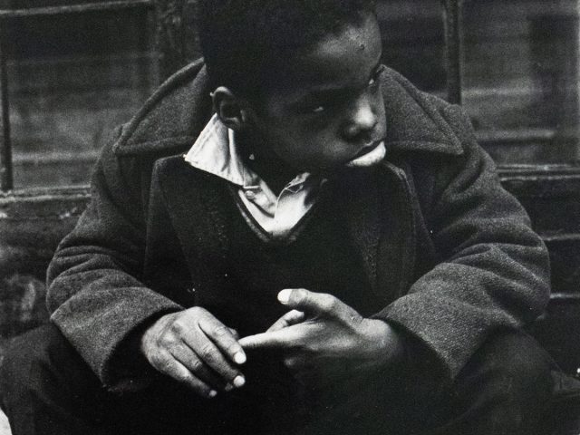A black and white photograph captures a young boy sitting on a concrete stoop, looking towards the right side of the frame. He wears a dark coat over a collared shirt and v-neck sweater, with his hands clasped.