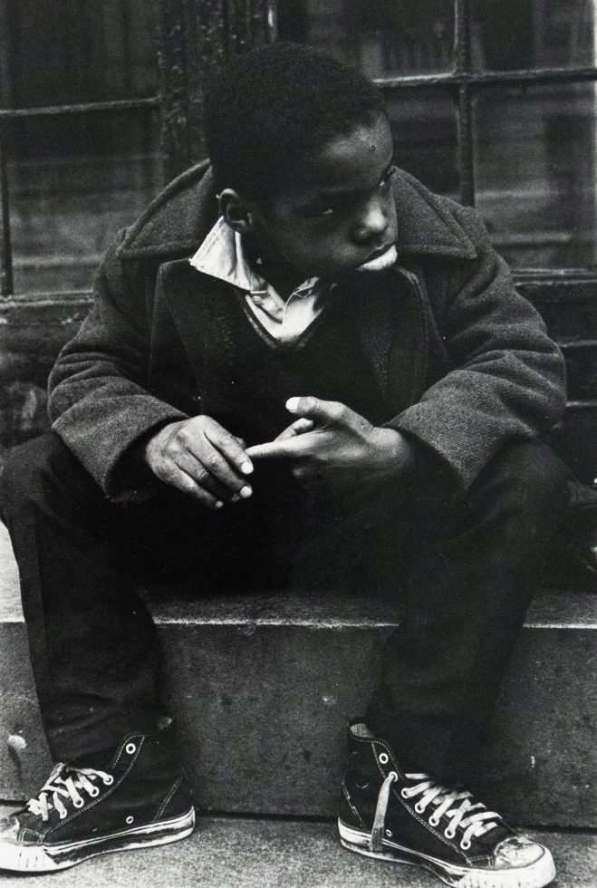 A black and white photograph captures a young boy sitting on a concrete stoop, looking towards the right side of the frame. He wears a dark coat over a collared shirt and v-neck sweater, with his hands clasped.