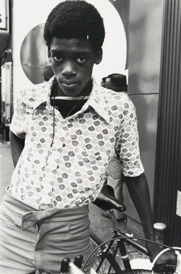 A black and white portrait shows a young boy with short, dark curly hair, leaning against a bicycle. He wears a patterned, short-sleeved shirt, light-colored pants, and a key on a string around his neck, gazing directly at the viewer with a serious expression.